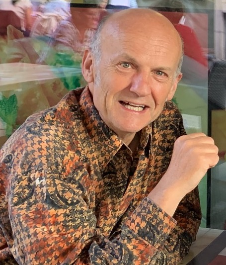 A white man with short grey hair wearing a plaid yellow shirt and smiling at the camera.